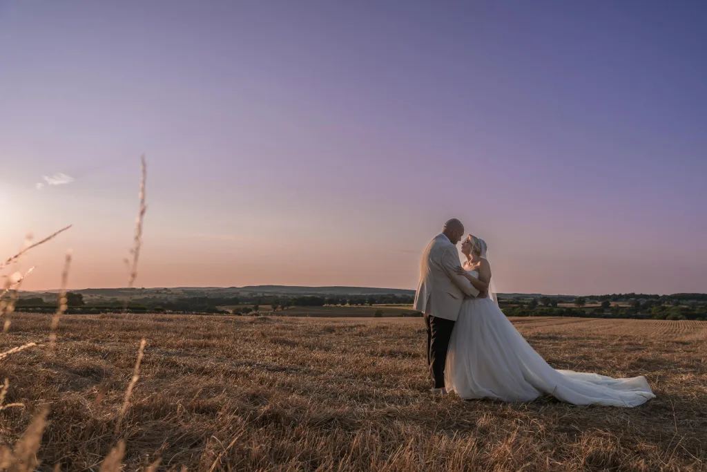 Bride and groom embracing in a golden field beneath a pink and lavender sunset sky.