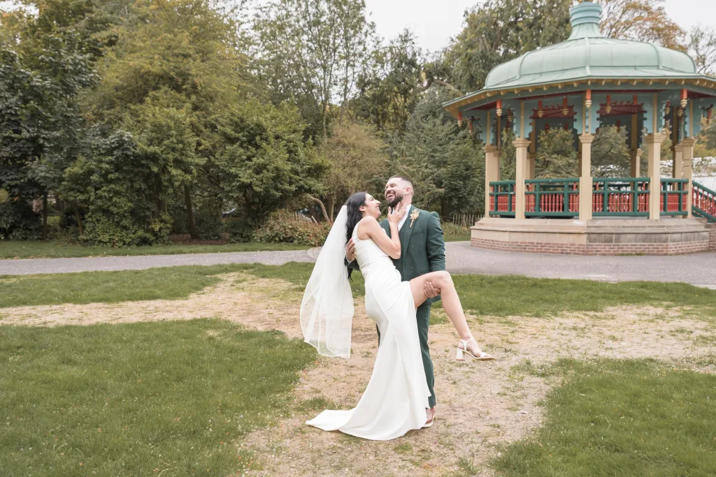 Bride laughing as her groom lifts her beside the bandstand in Pearson Park, Hull.