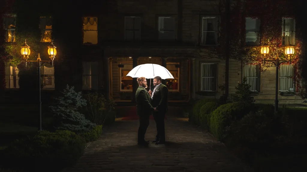 Two grooms beneath an illuminated umbrella during a nighttime wedding portrait.